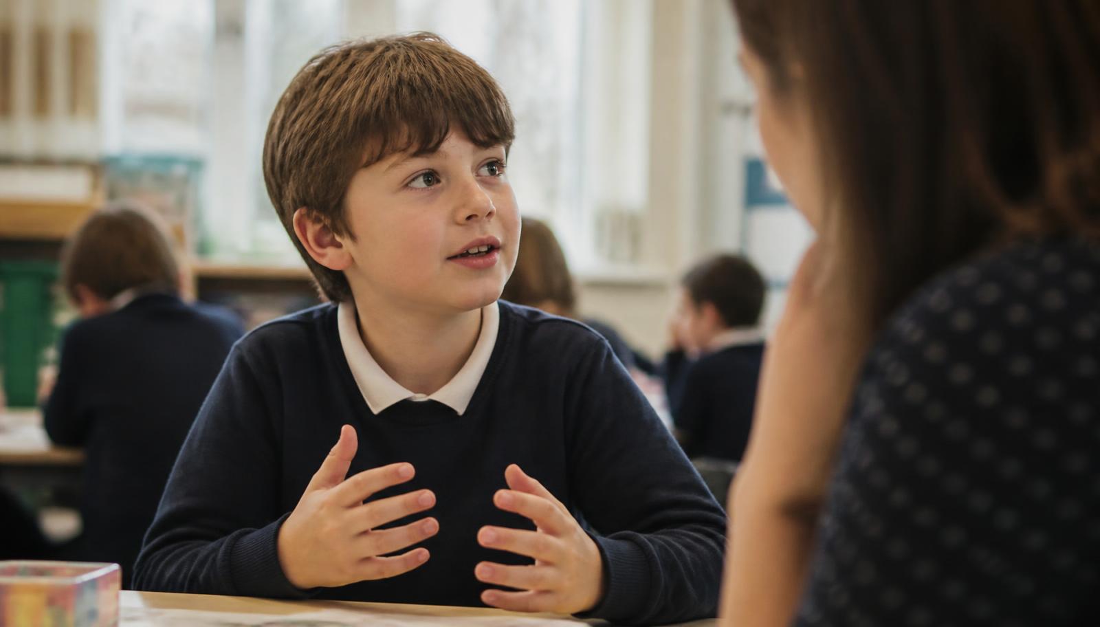 A young primary school child talking animatedly to an adult across a classroom table in soft window light.