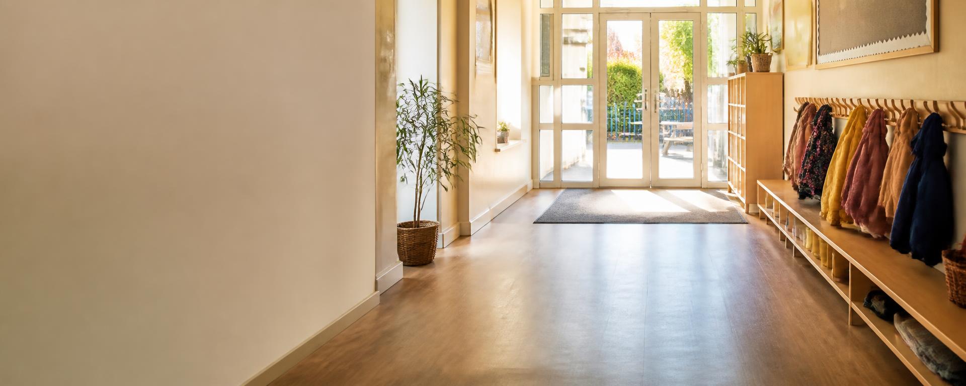 A sunlit primary school entrance hall with coats on low pegs and morning light through the doors.