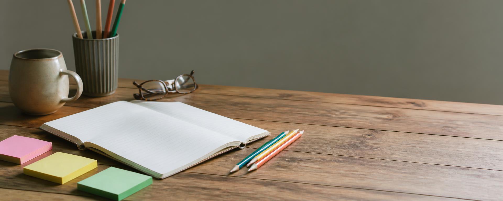 A wooden desk with an open notebook, coloured pencils and sticky notes in natural light.