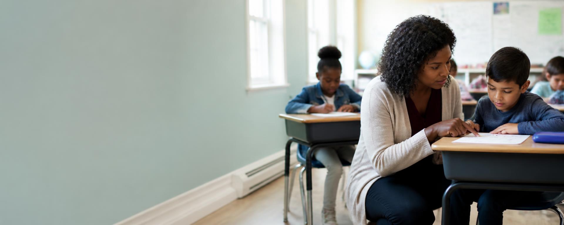 A teacher crouching beside a child's desk in a bright primary classroom, pointing at their work.
