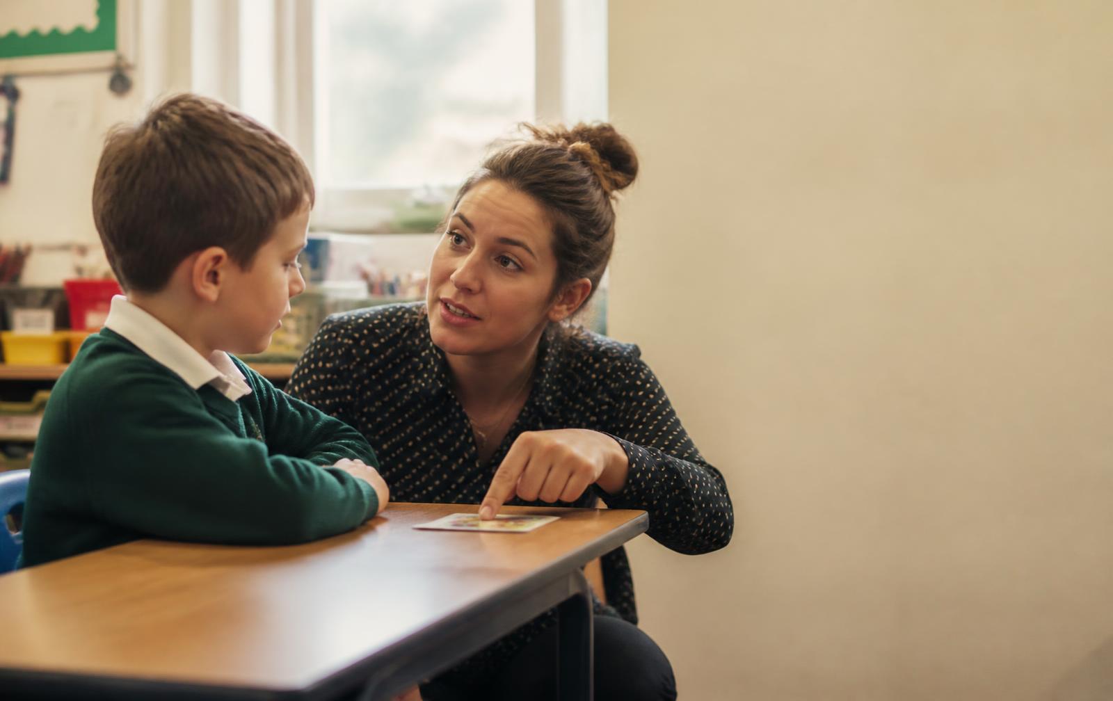 A teacher crouched at eye level beside a young child at a classroom desk, pointing at a small visual card in soft window light.