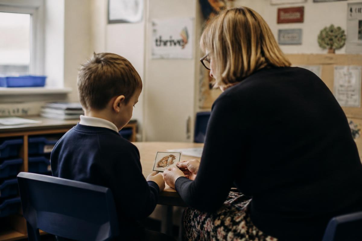 An adult and a young primary school child sitting side by side at a classroom table, looking together at a small picture card in soft window light.